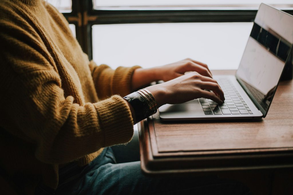Image of a person at a laptop with hands on keyboard. The laptop is sitting on a wood desk. The person is meant to be reviewing lines flagged by AI of responses that may be low quality.