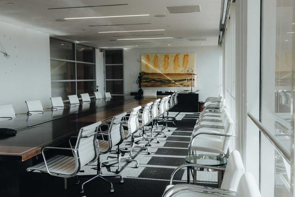 an empty conference room with a painting hanging on the far wall. The room is being viewed from a back right corner, and there are 9 white rolling chairs on each side of a long wooden table. There are more white chairs along the right side of the room, against a wall of windows. 